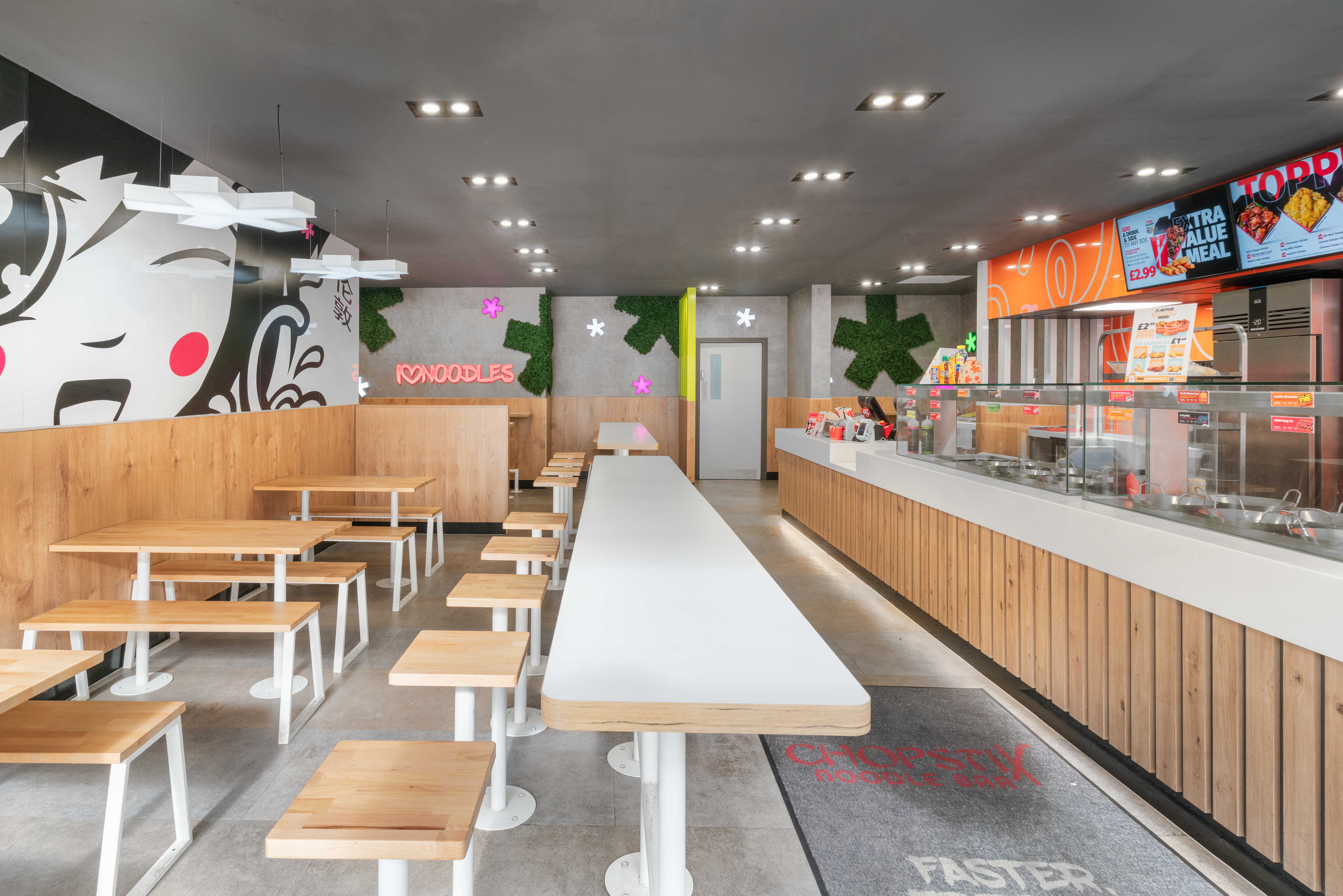 Modern noodle bar interior with long communal seating, wooden benches, branded wall graphics, and open service counter with digital menu boards.